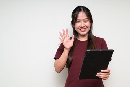 Studio portrait of young cute Asian woman against white background wearing casual t-shirt and using digital tablet computer for video call meeting while waving handの写真素材