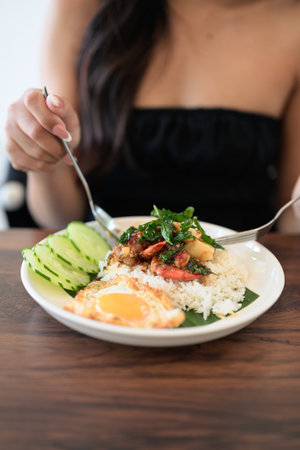 Close-up of woman in restaurant eating Thai holy basil seafood dish with fried egg and rice in restaurant during dayの写真素材