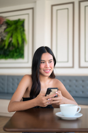 Beautiful young Asian woman sitting in cafe using mobile phone while having coffee cup on table during day remote work conceptの写真素材