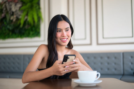 Beautiful young Asian woman sitting in cafe using mobile phone while having coffee cup on table...