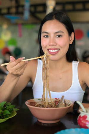 Beautiful young Thai woman eating famous Thailand street food boat noodles served at local...