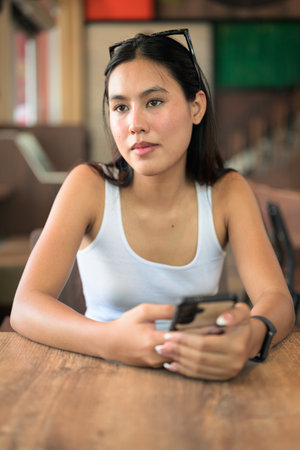 Beautiful young Asian woman sitting at restaurant using mobile phone texting and thinkingの写真素材