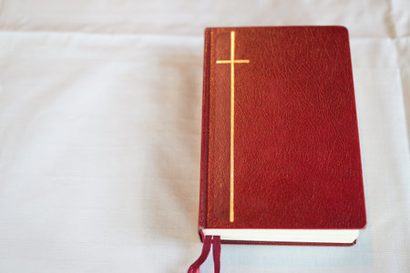 Red holy bible on a white bed with cross in the background.