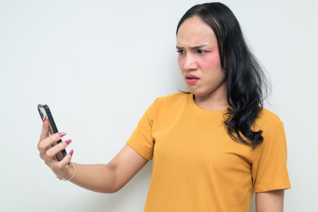 Studio portrait of a young Thai transgender individual with makeup and long hair using phone. Concept of gender expression, LGBTQ pride, and inclusivity.の写真素材