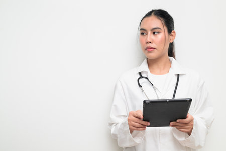 Portrait of a young Thai female doctor in a white medical uniform using a digital tablet computer with a stethoscope on plain background, suitable for healthcare advertising or medical communication.の写真素材