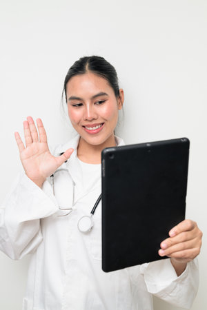 Portrait of a young Thai female doctor in a white medical uniform using a digital tablet computer with a stethoscope on plain background, suitable for healthcare advertising or medical communication.の写真素材