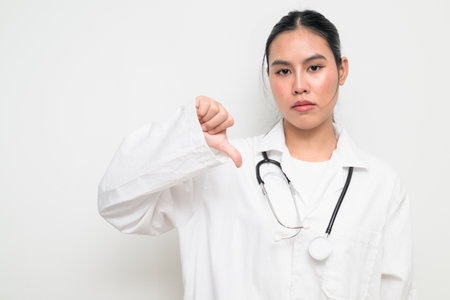 Portrait of a female healthcare professional in a white medical uniform with a stethoscope, standing against a clean studio background giving thumbs down gesture.の写真素材