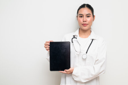 Portrait of a young Thai female doctor in a white medical uniform using a digital tablet computer with a stethoscope on plain background, suitable for healthcare advertising or medical communication.の写真素材