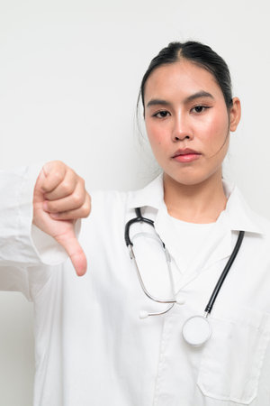 Portrait of a female healthcare professional in a white medical uniform with a stethoscope, standing against a clean studio background giving thumbs down gesture.の写真素材