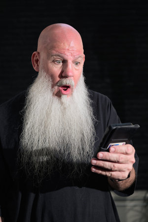 Portrait of old bald man with a long white beard using phone against a dark backgroundの写真素材