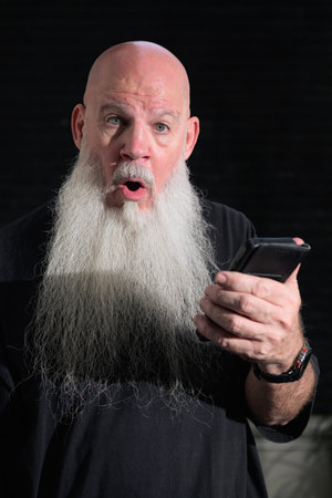 Portrait of old bald man with a long white beard using phone against a dark background