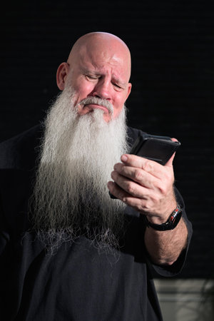 Portrait of old bald man with a long white beard using phone against a dark background