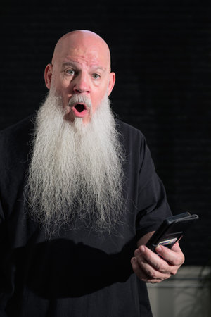 Portrait of old bald man with a long white beard using phone against a dark background