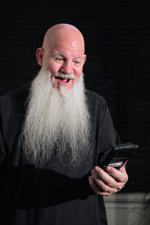 Portrait of old bald man with a long white beard using phone against a dark background