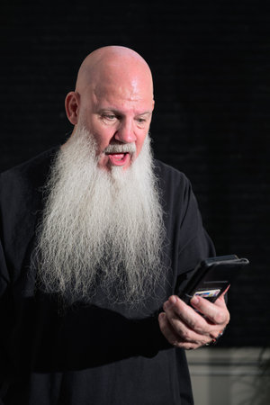 Portrait of old bald man with a long white beard using phone against a dark background