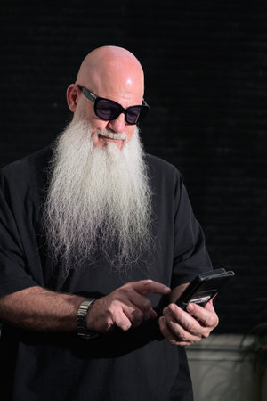 Portrait of a bald man with a long white beard and sunglasses with his smartphone outdoors