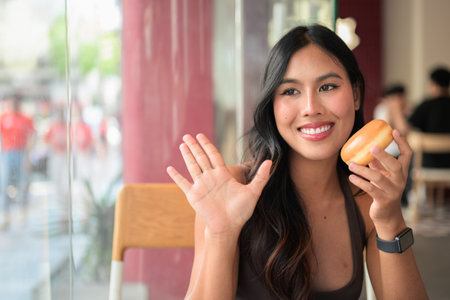 Young Asian woman sitting in a cafe holding a glazed donut while waving her hand in greeting