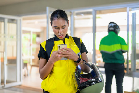 A focused young Asian female delivery driver looks down at her smartphone while holding a motorcycle helmet under her arm. She wears a yellow high-visibility vest and a smartwatch, standing outside a glass office doorway with another courier blurred in the background.