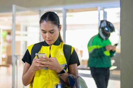 A horizontal medium shot of a focused young Asian female delivery driver looking at her mobile phone screen. She wears a yellow high-visibility safety vest and stands outdoors under a covered building entrance while a blurred male coworker in a green jacket operates his phone in the background.