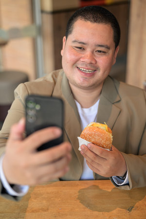 A happy plus size mid adult Asian businessman from the Isan region smiles while taking a mobile phone selfie with a hamburger at a restaurant table with a shallow depth of field.