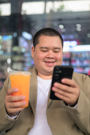A happy plus size young adult Asian businessman from the Isan region sits outdoors smiling while using a smartphone and holding an orange juice drink at a night cafe.