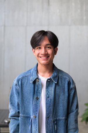 Vertical lifestyle photograph of a handsome young Southeast Asian Thai Isan non-binary man smiling happily and looking directly at the camera while standing against a corrugated metal wall with copy space.