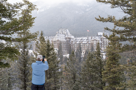 Banff, Alberta Canada - Jan 21, 2019: Male tourist photographs a  winter scene view of the famous Fairmont Banff Springs Hotel, as seen from Surprise Corner in Banff National Park with a smartphoneのeditorial素材