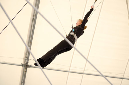 An adult female hangs on a flying trapeze at an indoor gym. The woman is an amateur trapeze artist.