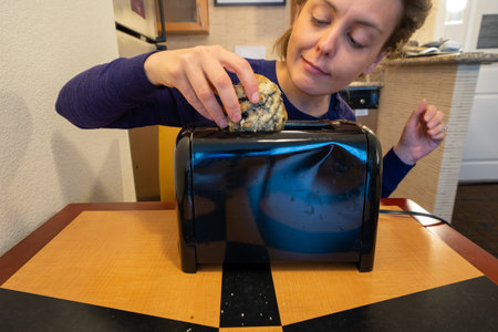 Misguided woman attempts to stuff a large blueberry scone into a small toaster slot, meant for bread