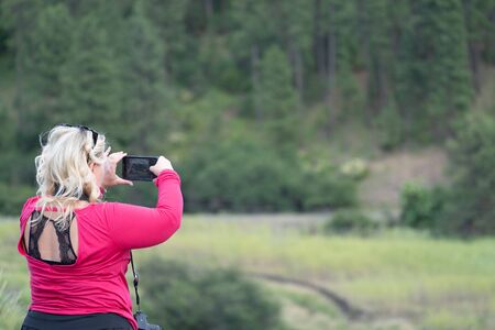 Blonde woman photographer takes pictures with her smart phone at a scenic overlookの写真素材