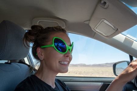 Young adult woman wearing alien sunglasses drives in a car along Nevada's Extraterrestrial Highway....