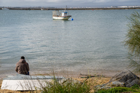 Isla Cristina, Spain - January 23, 2020: Young man sits on the beach, watching boats in the marshlandの写真素材