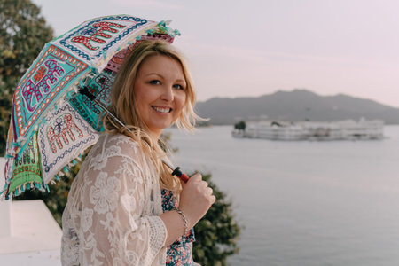 Smiling blond woman wearing a lace top holding a parasol umbrella overlooking Pichola Lake in Udaipur Indiaの写真素材