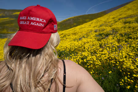 Taft, California - March 25, 2019: Blonde woman wearing a Donald Trump Make America Great Again hat, sitting in a field of yellow wildflowers. Concept for the 2020 United States Presidential Electionのeditorial素材