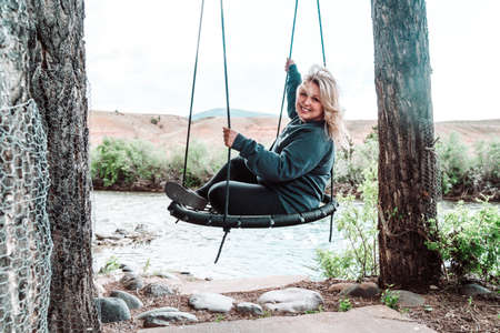 Happy, relaxed blonde woman sits on a tree swing near a river, looking happyの写真素材