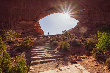 Tourists (unidentifiable) stand in the middle of the North Arch in the Windows Section of Arches National Park in Utah, with sunflare at sunriseの写真素材