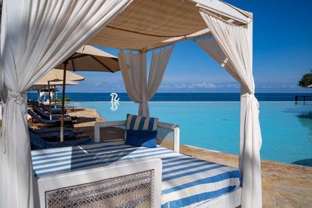 Zanzibar, Tanzania - March 16, 2023: Logo with an R for the Royal Zanzibar Resort, in front of the infinity swimming pool and the Indian Ocean and beach cabanasのeditorial素材