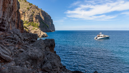A sleek white yacht is anchored in the deep blue waters near the rugged coastline of Sa Calobra on the island of Mallorca, Spain, under a clear summer sky.の写真素材