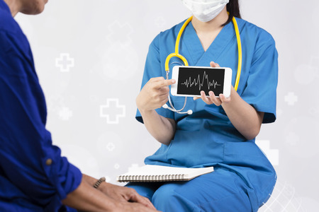 Female doctor showing heart rate monitor application on mobile to her elderly patient over abstract white background with hospital icon.の写真素材