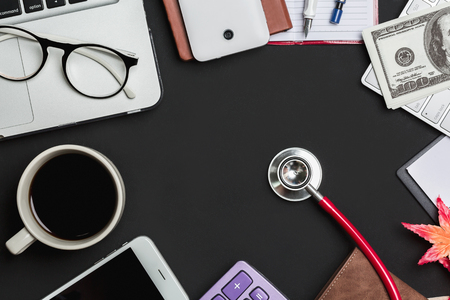 Desk table of doctor with laptop, coffee cup, stethoscope with a lot of things on black, view from above.の写真素材
