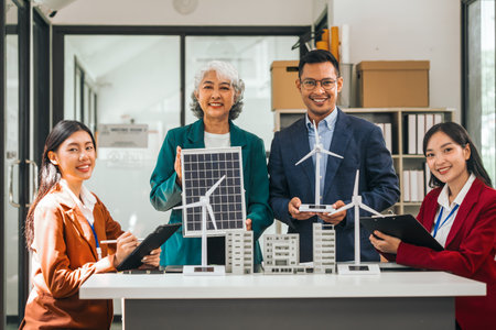 Four asian businesspeople professionals discuss renewable energy in office, showing solar panel and wind turbines with tower building model, apartment. modern business with environmental, clean energyの写真素材
