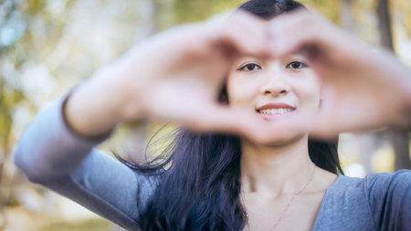 Young woman outdoors forming a heart shape with hands in front of smiling face, soft focus, autumn...