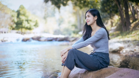 Young woman sitting on a riverside rock, smiling and relaxing in nature with soft morning light and...