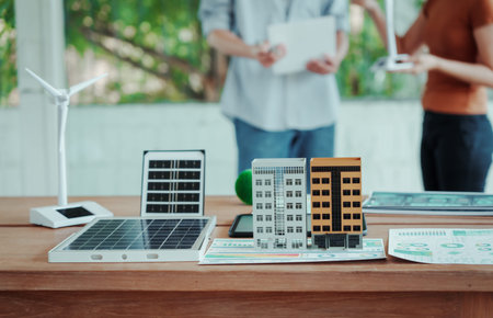 Tabletop model of solar panels, wind turbine and miniature buildings for renewable energy planning; blurred people discussing in background, modern office setting.