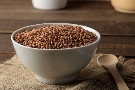 Dry buckwheat groats in a white bowl with a spoon on a wooden brown table. cereals. healthy food. porridge.の写真素材