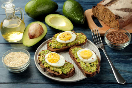 Sandwich with avocado puree and egg on a plate and ingredients for cooking on a blue wooden table.の写真素材