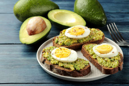 Sandwich with avocado puree and egg on a plate and ingredients for cooking on a blue wooden table.の写真素材