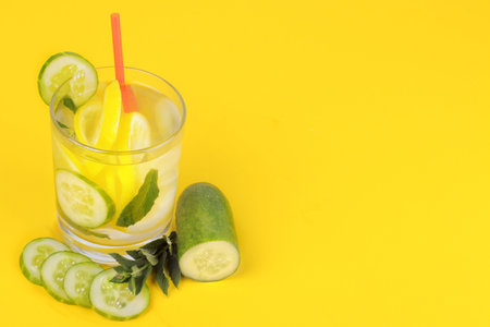 Refreshing water with lemon and cucumber and mint in a glass cup next to a fresh cucumber on a bright yellow paper backgroundの写真素材