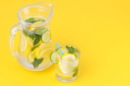 Refreshing water with lemon and cucumber and mint in a glass cup and jug on a bright yellow paper...