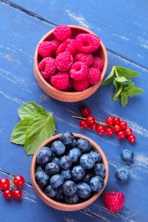 Fresh, large blueberries and raspberries in a wooden bowl close-up with mint stamens on a blue wooden background. top viewの写真素材